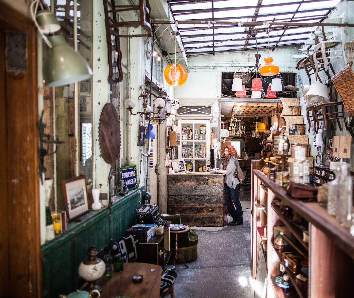 entrée de l'Etablisienne depuis la rue avec Laurence Sourisseau, fondatrice du lieu, adossée au comptoir d'accueil. Divers objets sont disposés un peu partout au sol et sur des meubles.