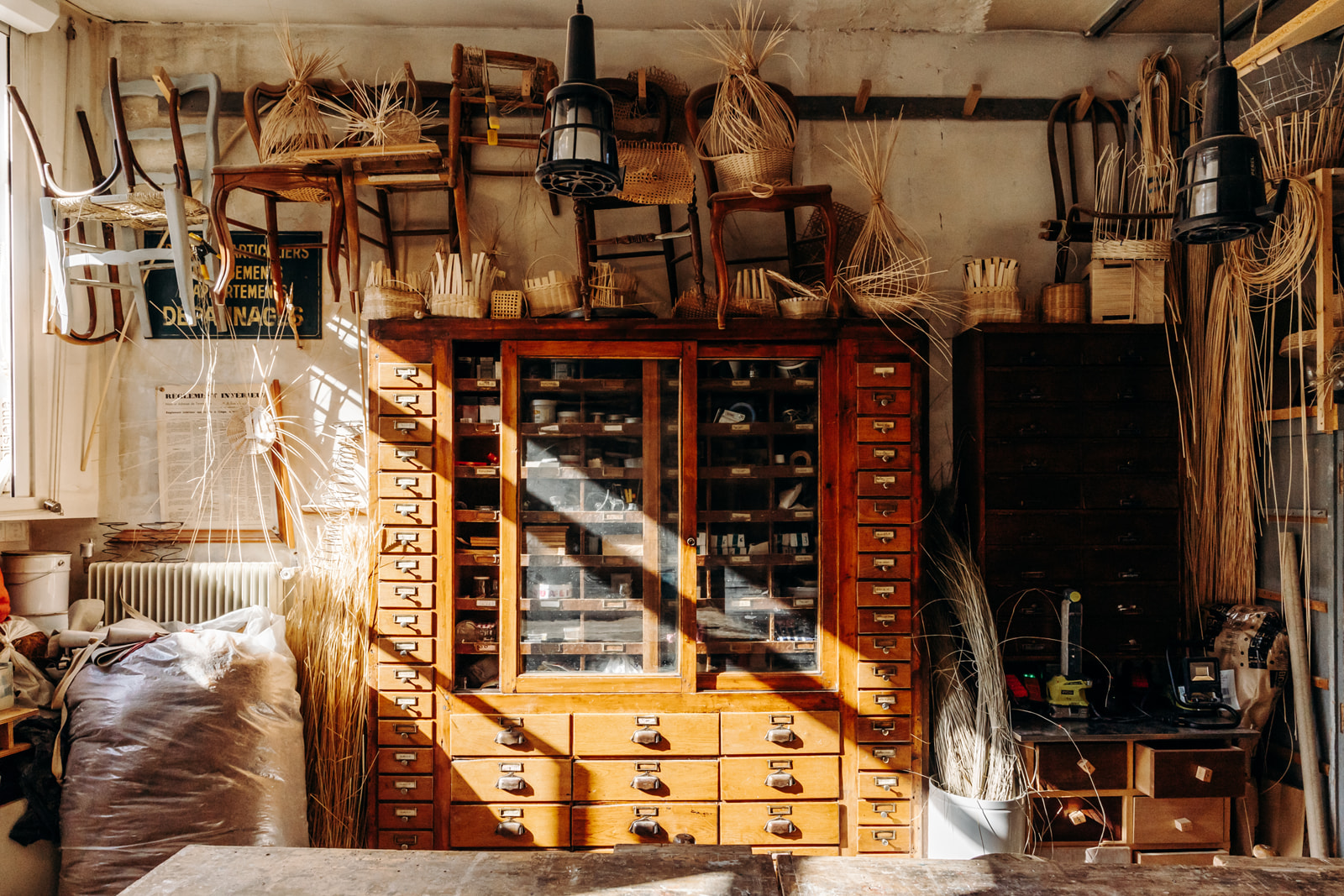 vue de l'atelier du rez-de-chaussée avec une grande table en bois au centre de la pièce, une grande armoire à tiroirs et des outils et machines rangés sur des meubles